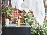 Moderne køkkenvask med sort bordplade, vask i rustfrit stål, hvid sæbedispenser, grønne planter, murstensvæg og hængende håndklæder.