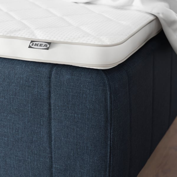 En seng med en hvid madras på en mørk bund, der fremhæver forstærkede kanter for stabilitet, med en IKEA-etiket.