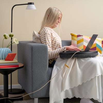 Kvinde i stribet skjorte arbejder på laptop ved bordet med lampe og blomster.