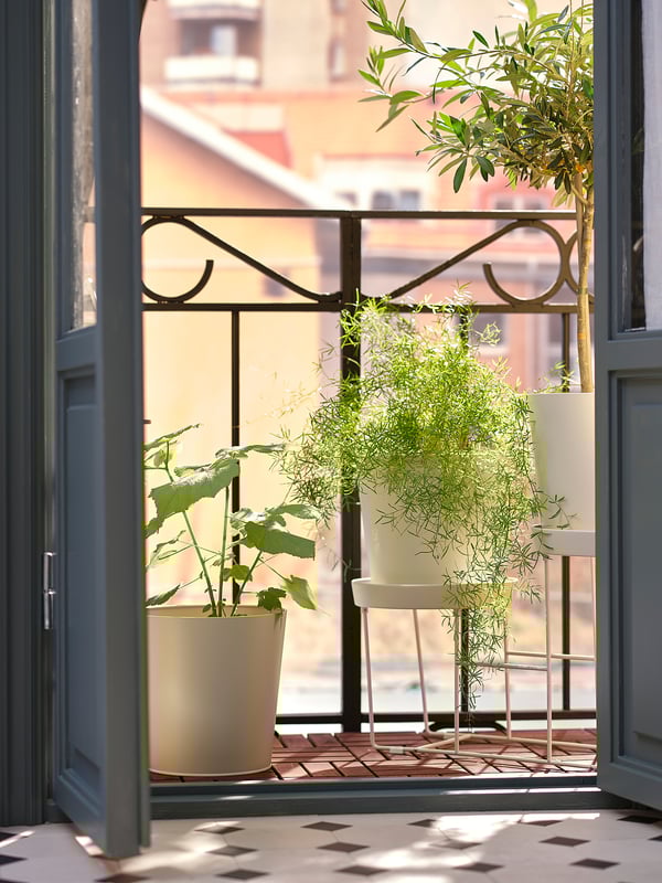 En balkon med RUNNEN terrasse i beige, med et hvidt metalrækværk. Tre potteplanter forstærker den varme, indbydende solrige atmosfære.