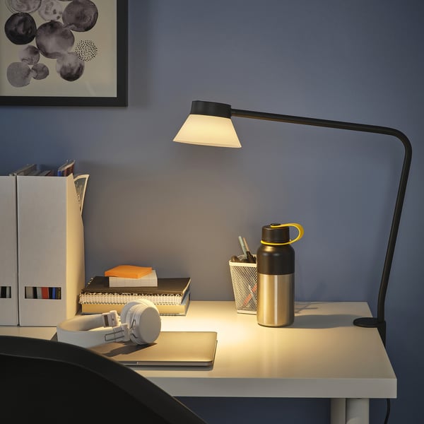 SAMBORD-lampe med trekantet skærm med skrivebordsklemme, der oplyser arbejdsplads med bøger, flaske og hovedtelefoner.