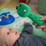 Baby holder grøn frørangle, smilende, med blå ugle legetøj ved siden af sig.