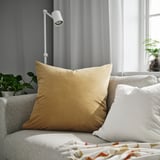 Stor guld SANELA pude på beige sofa ved siden af hvid pude, moderne lampe, grøn plante og seng med farverigt tæppe.