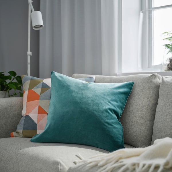 Moderne stue med en grå sofa, en stor turkis SANELA fløjlspude og en mønstret pude mod et vindue med lyse gardiner.