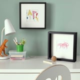 Et skrivebord med en grøn kop, kontorartikler, to indrammede billeder, lampe og dinosaurlegetøj.