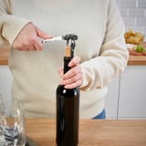Person åbner vinflaske med proptrækker. Scene på træbord med glas.
