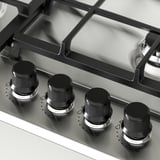 Fire knapper på et gaskomfur, sort med metallisk bund, justeret på et sølv kontrolpanel, hvilket indikerer design og funktionalitet af høj kvalitet.