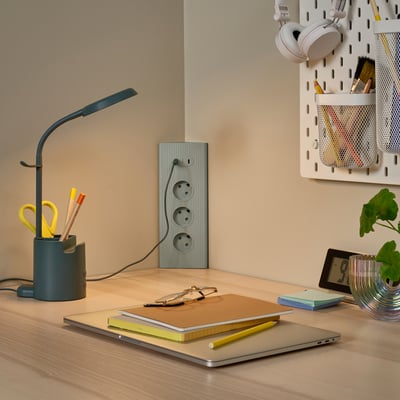 Træskrivebord med grøn SKOPAT-ledning, bærbar computer, notesbøger, hovedtelefoner og en plante.