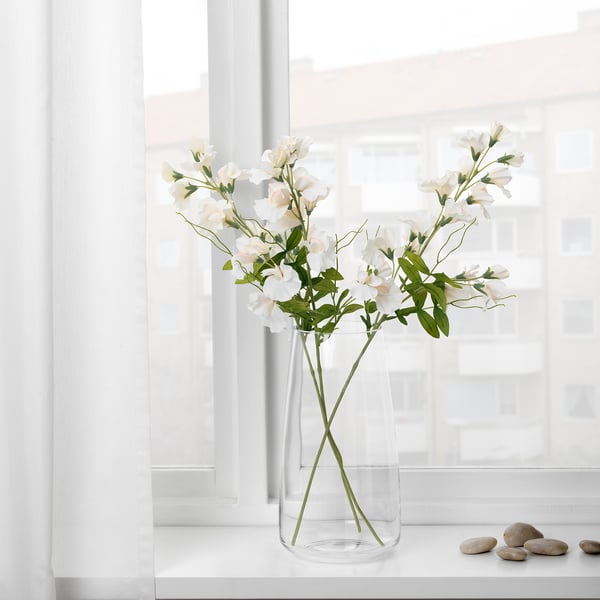Vase med kunstige hvide blomster ved vindue. Slankt klart glas holder bøjelige, ståltrådede stængler, der reflekterer naturligt lys.