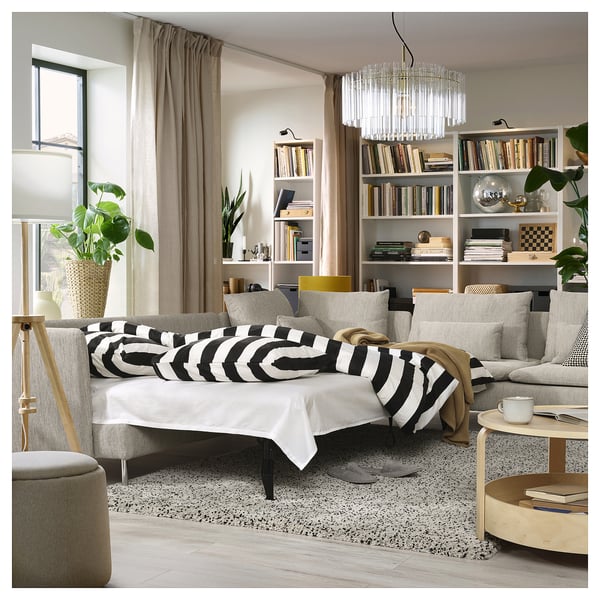 Beige sovesofa, stribede puder, træbord, lysekrone, bogreol med bøger og planter, stort vindue med gardiner.