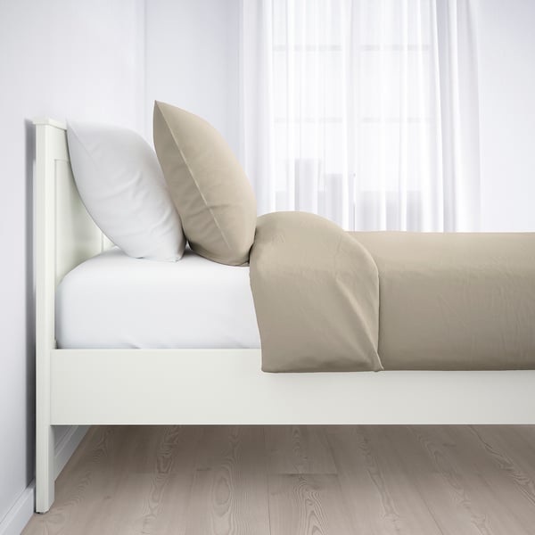 Minimalistisk hvid seng med beige puder, sat op mod gennemsigtige hvide gardiner på et trægulv.