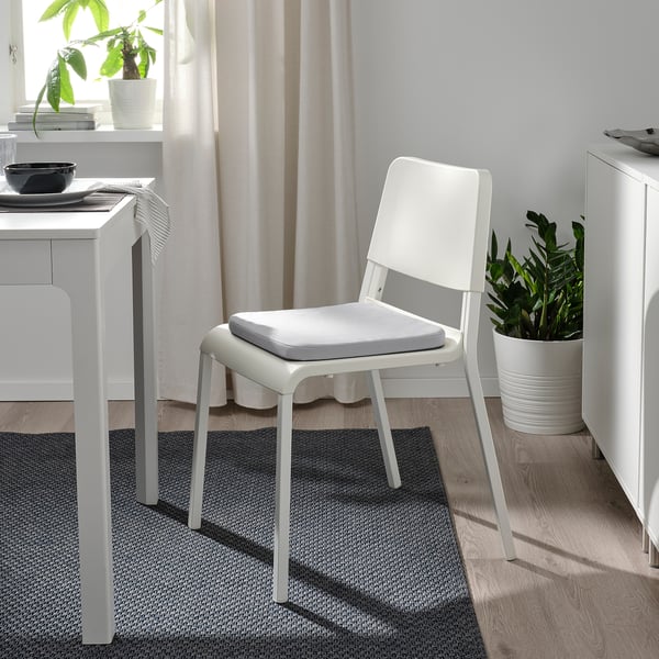 Minimalistisk spiseplads: hvid staggastrarr stol, grå hynde, nær bord og plante. Lysfyldt.