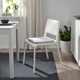 Minimalistisk spiseplads: hvid staggastrarr stol, grå hynde, nær bord og plante. Lysfyldt.