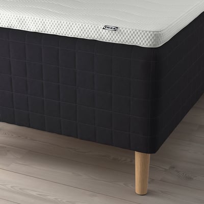 Nærbillede af en seng med mørk madras, hvid topmadras og træben, der viser et IKEA-mærke.