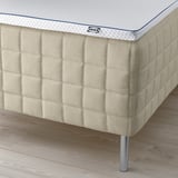 En beige seng med en tekstureret madras, toppet med et blåt IKEA tag. Den har metalben til støtte.