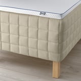 Et hjørne af en IKEA STUVLAND madras på et trægulv. Den viser en madras med linnedtekstur, understøttet af synlige træben.