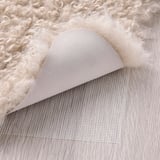 Fluffy, cremefarvet tæppe, bagside af beige stof og skridsikkert mesh, perfekt til varme og komfort.