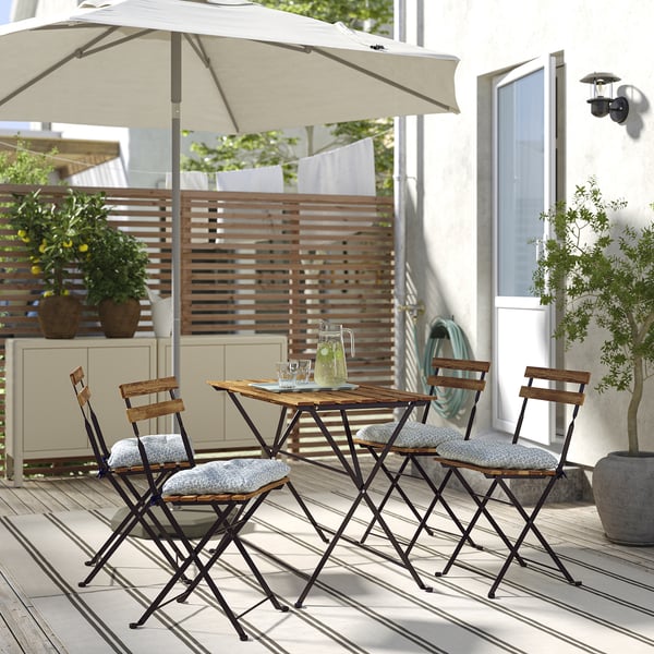 Lille udendørs spisebordssæt, TÄRNÖ, med sammenklappeligt træbord og 4 stole, lyse hynder, under en stor parasol på en terrasse.