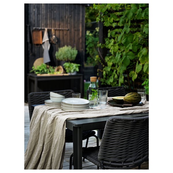 Udendørs terrassesæt med sort bord og stole, hørdug, tallerkener, glas og vandmelon på bordet. Omgivet af grønne områder og træhegn.