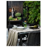 Udendørs terrassesæt med sort bord og stole, hørdug, tallerkener, glas og vandmelon på bordet. Omgivet af grønne områder og træhegn.