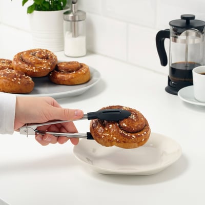 En hånd bruger en tang til at lægge et wienerbrød på en hvid tallerken, med flere kager og en kaffepresse i baggrunden.