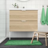 Moderne badeværelse med lyst træ forfængelighed, hvid bordplade og vask. Grøn TOFTBO måtte, håndklæder og bænk er pænt arrangeret. Måtten er lille.