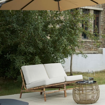 Moderne flet udendørs loveseat med hvide hynder og rundt vævet sidebord under stor solbrun paraply ved siden af frodige grønne buske.
