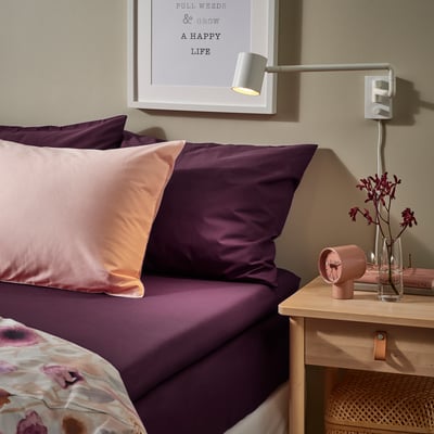 Seng med lilla ULLVIDE-lagner, lyserøde puder. Vægkunst, moderne lampe, blomster, bøger på natbord. Hyggelig, stilfuld indretning.