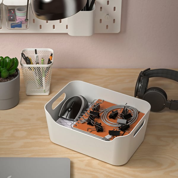 Træskrivebord med organiserede kontorartikler. Hvid skraldespand rummer små genstande som clips og snore. Hovedtelefoner og en lukket bærbar computer er også til stede.