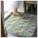 Person, der træder på VÄLFÄRD-måtten, farverigt blomsterdesign, polyester- og gummimaterialer, placeret ved en indgang.