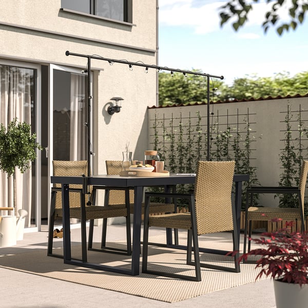 Moderne terrassesæt med rektangulært sort metalbord, fire kurvestole og hængende lyskæder, ideel til udendørs spisning eller afslapning.