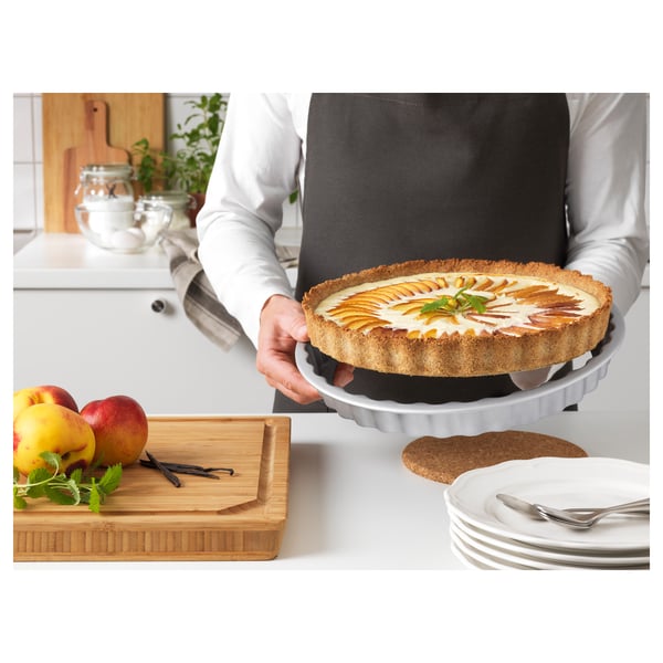 Person, der holder en friskbagt tærte i en grå non-stick tærteform, klar til servering. På et bord er der frugt, tallerkener og redskaber.