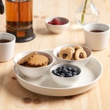 Et køkkenbord med småkager, bær og kaffe i kopper omkring en roterende drejeskive med flere snacks.