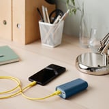 Skrivebord med telefonopladning fra blå powerbank, gult kabel, metallisk lampe, grøn notesblok, blyantholder og glas.