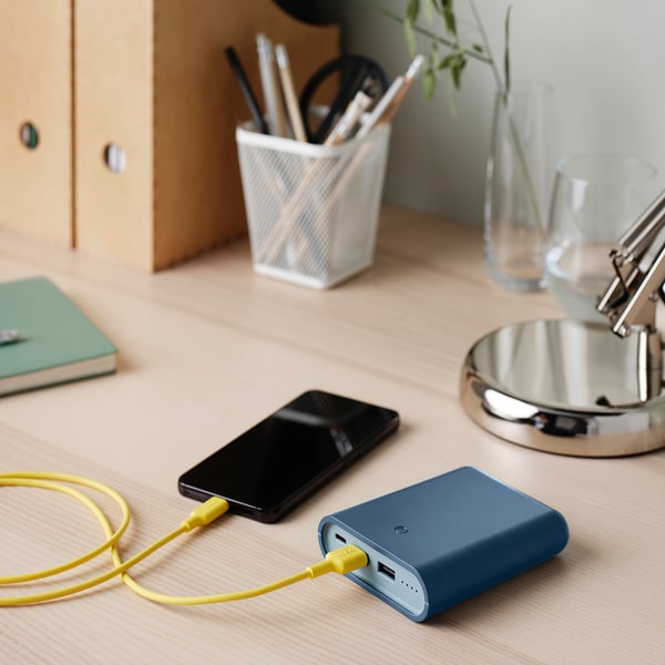 Blå powerbank på skrivebordet, oplader telefon med gult kabel. Mindre end kop, ved siden af kuglepen, grøn notesbog og lampe.