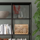 Sort hylde med glas, vaser, røde blomster, vævet kurv, bøger, beige skål, plante.