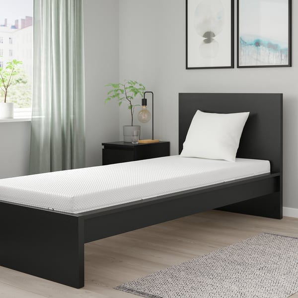 Modern bedroom with black mattress, pillow, bedside table, lamp, and plant against a grey wall with pictures.