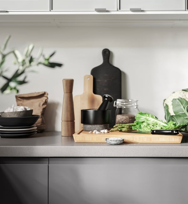 A kitchen counter with tools: black mortar, wooden pestle, knife, vegetables, bowls, and plates.