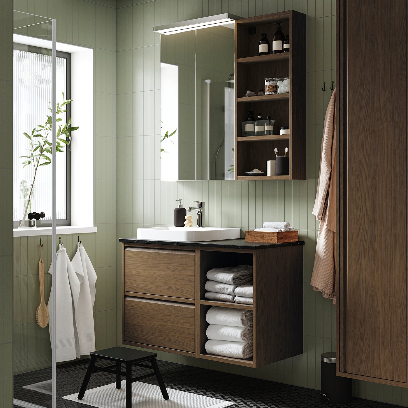 Modern green bathroom with wall-mounted brown oak cabinet, white sink, open shelves, and hanging towels. Mirror and plant by window.