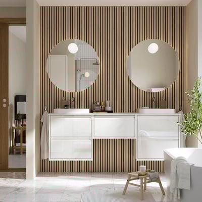 Modern bathroom with white ÄNGSJÖN vanity, large round mirrors, marble-like countertop, and wooden stool. Vertical wooden slat backsplash.