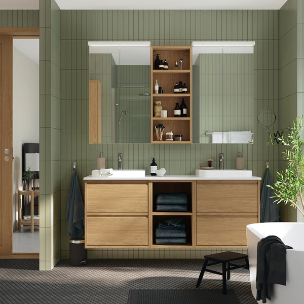 Modern bathroom with green walls, wooden cabinets, dual sinks, and open shelves. A small stool rests near the tub.