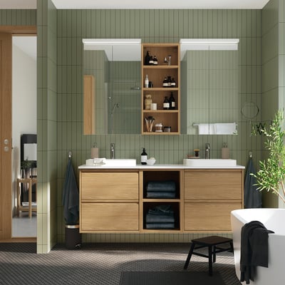 Modern bathroom with green walls, wooden cabinets, dual sinks, and open shelves. A small stool rests near the tub.
