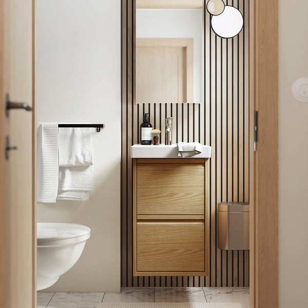 Modern bathroom with oak cabinet, sleek counter, minimal handles. White sink, towels, black accents. Ample storage.