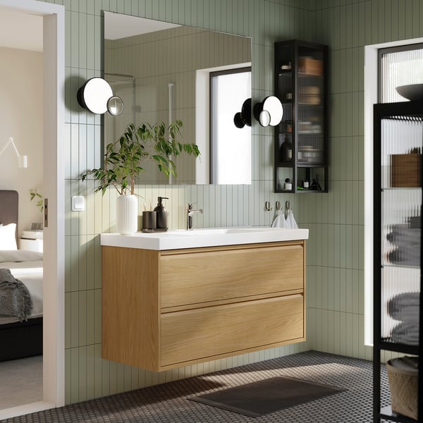 Modern bathroom with oak vanity, mirror, plants, and glass shelves. ÄNGSJÖN series blends wood and minimalist design.