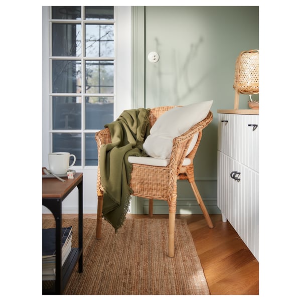 A cosy corner with a woven wicker chair, green throw, white pillow, and side table with a mug.