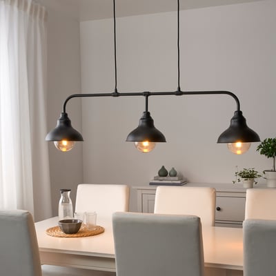 An AGUNNARYD ceiling lamp with a lengthy 122cm black metal rail and three dark lampshades glowing warmly, hanging prominently above a dining table.