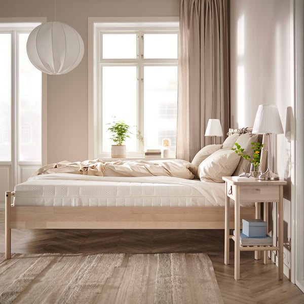 Modern minimalist bedroom with beige ÅKREHAMN bed, natural wooden floor, large windows, hanging lamps, and wooden bedside table.