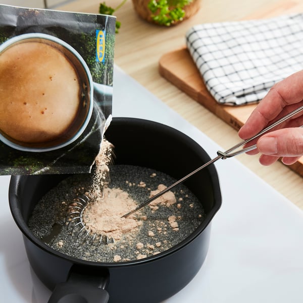 Person pours light brown powder into black bowl with metal spoon, wooden surface, cloth, plant.