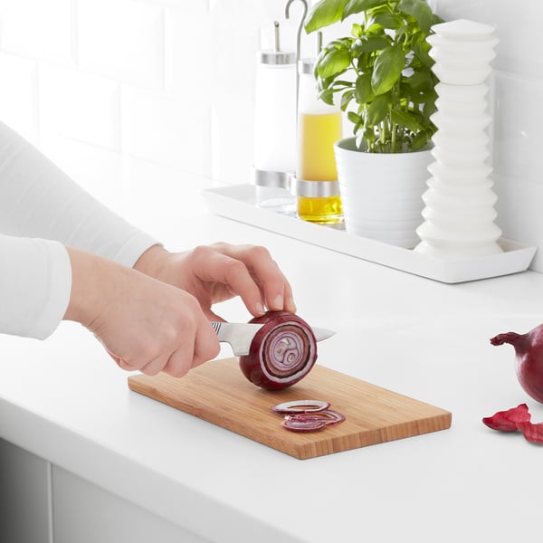 Person slices onion on bamboo chopping board on white countertop, with kitchen accessories behind.