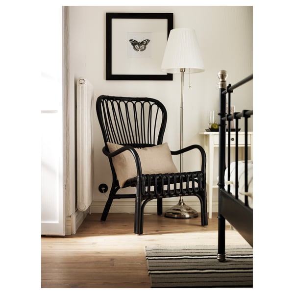 Black rattan-style armchair with beige cushion under black-framed butterfly art, next to a white lamp with a silver base on a wooden floor.
